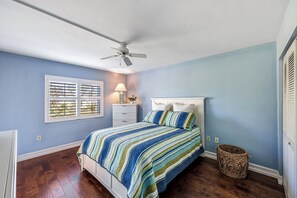 Un dormitorio con una cama, un ventilador de techo, una mesita de noche, una lámpara, una ventana con persianas y una cesta de mimbre.