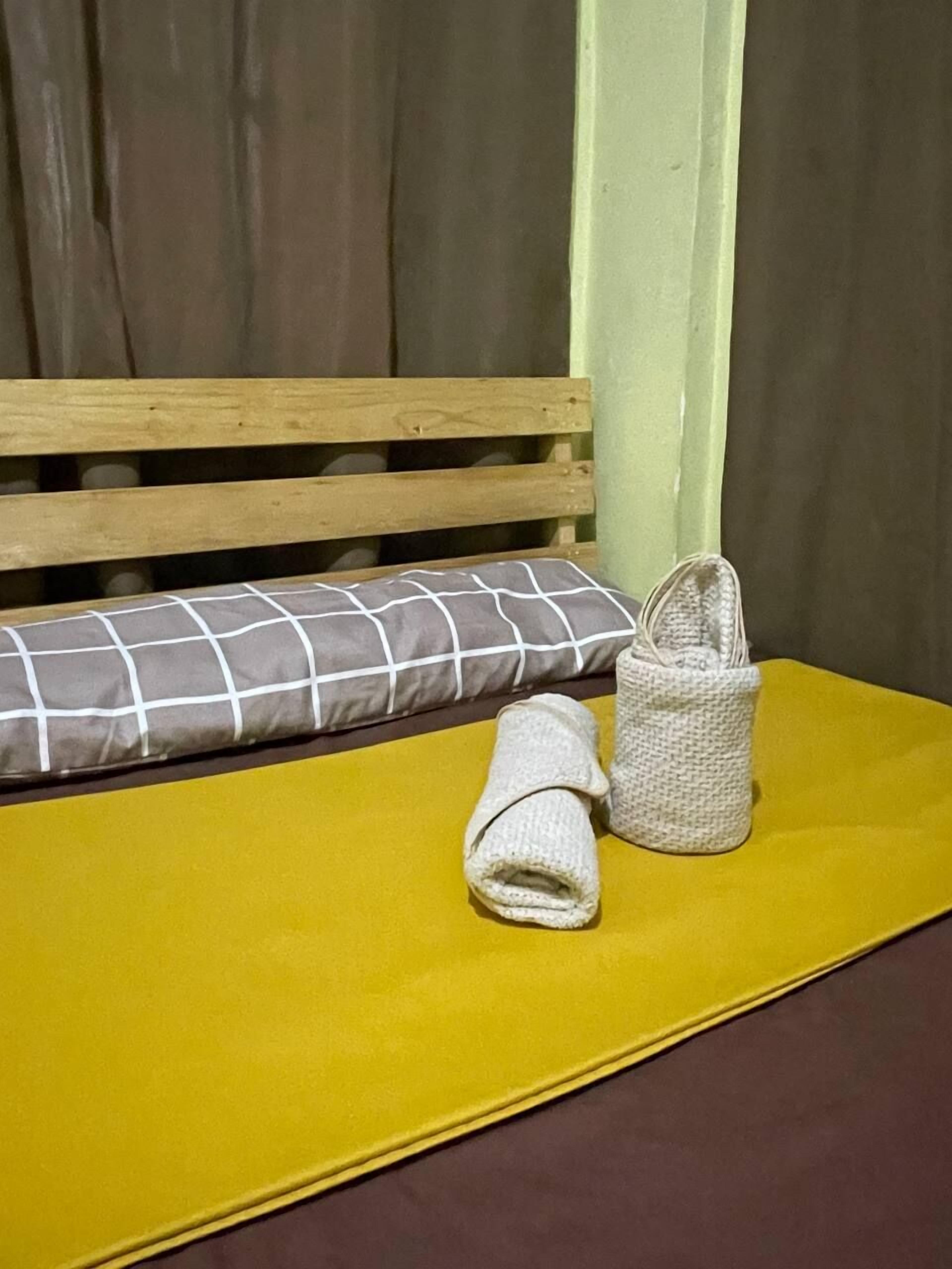 A bed with a wooden headboard, a yellow mattress, a grey and white checkered pillow, and two rolled towels.