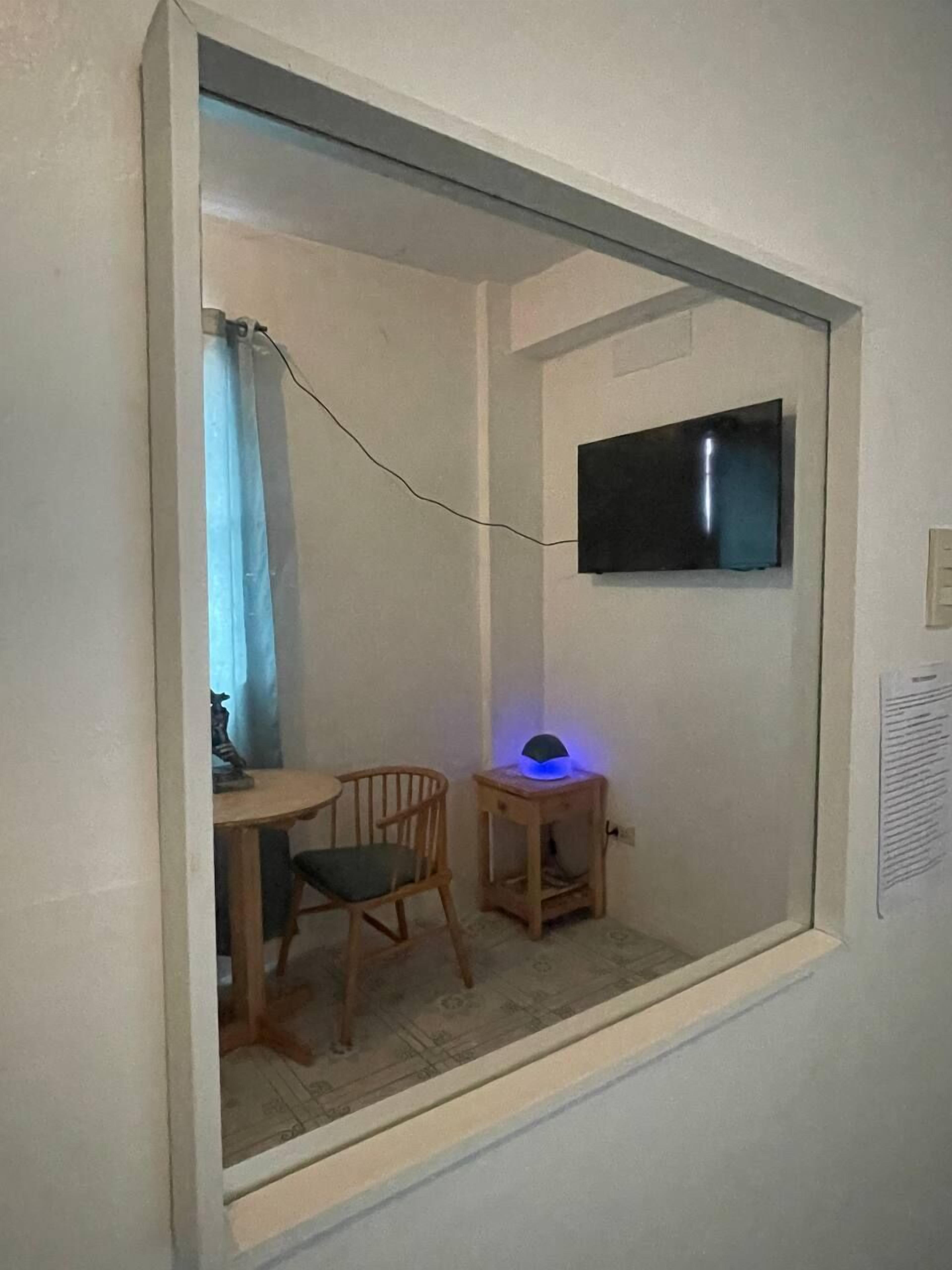 Una habitación pequeña con una televisión montada en la pared, una silla de madera, una mesita y una lámpara con luz azul.