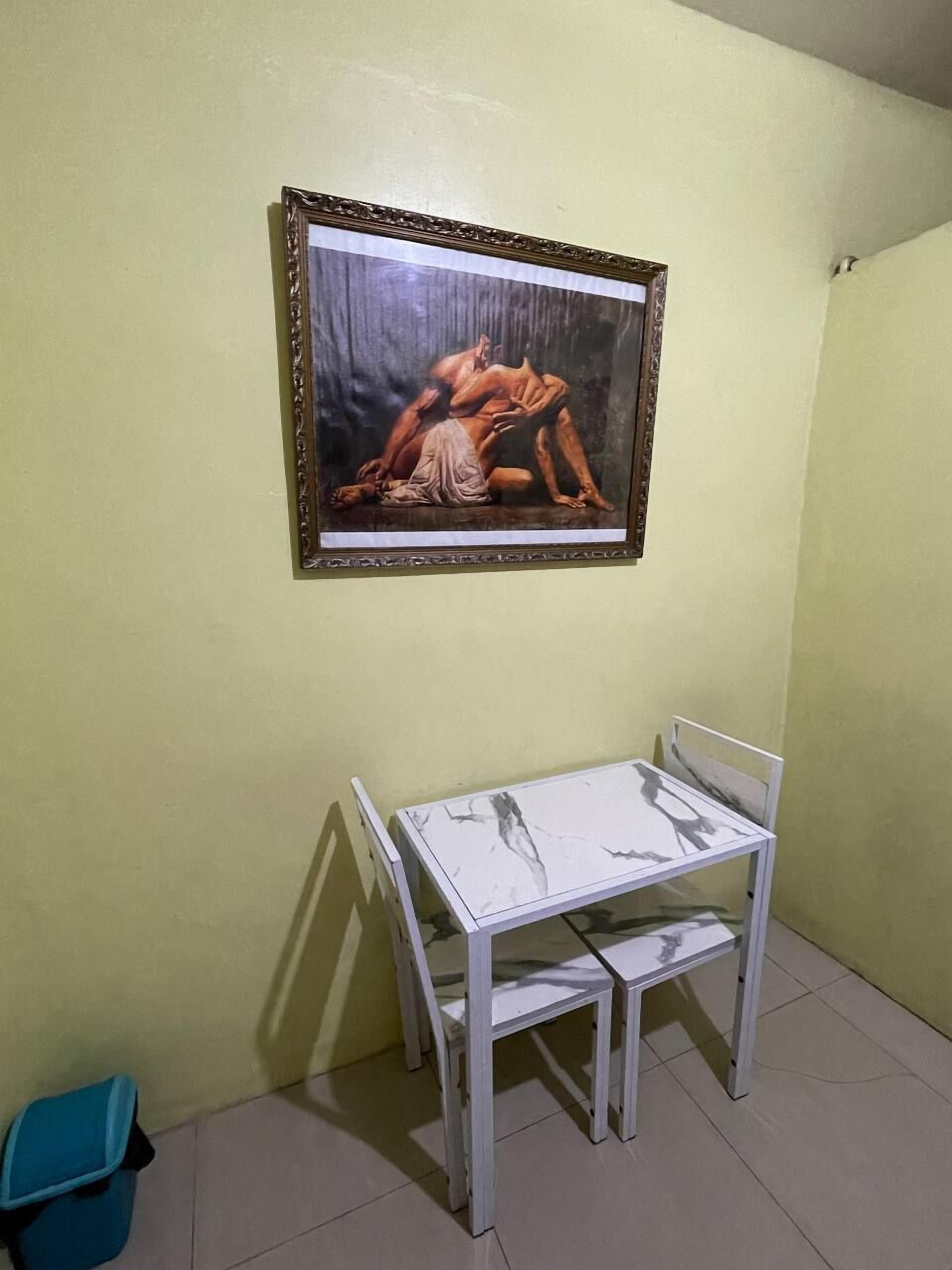 Quarto com uma pintura emoldurada de uma figura nua, uma mesa de metal branca e uma lixeira azul.