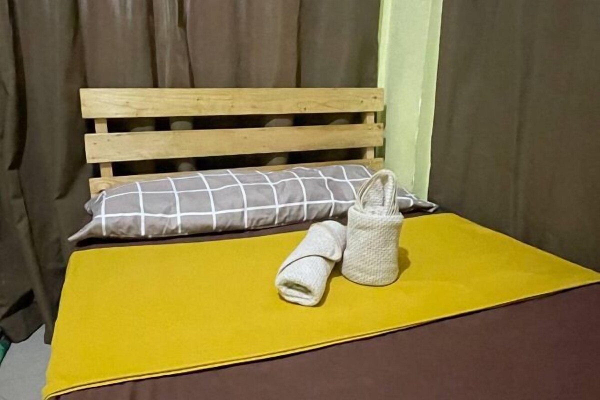 A bed with a wooden headboard, a yellow bedspread, a rolled towel, and a small woven basket.