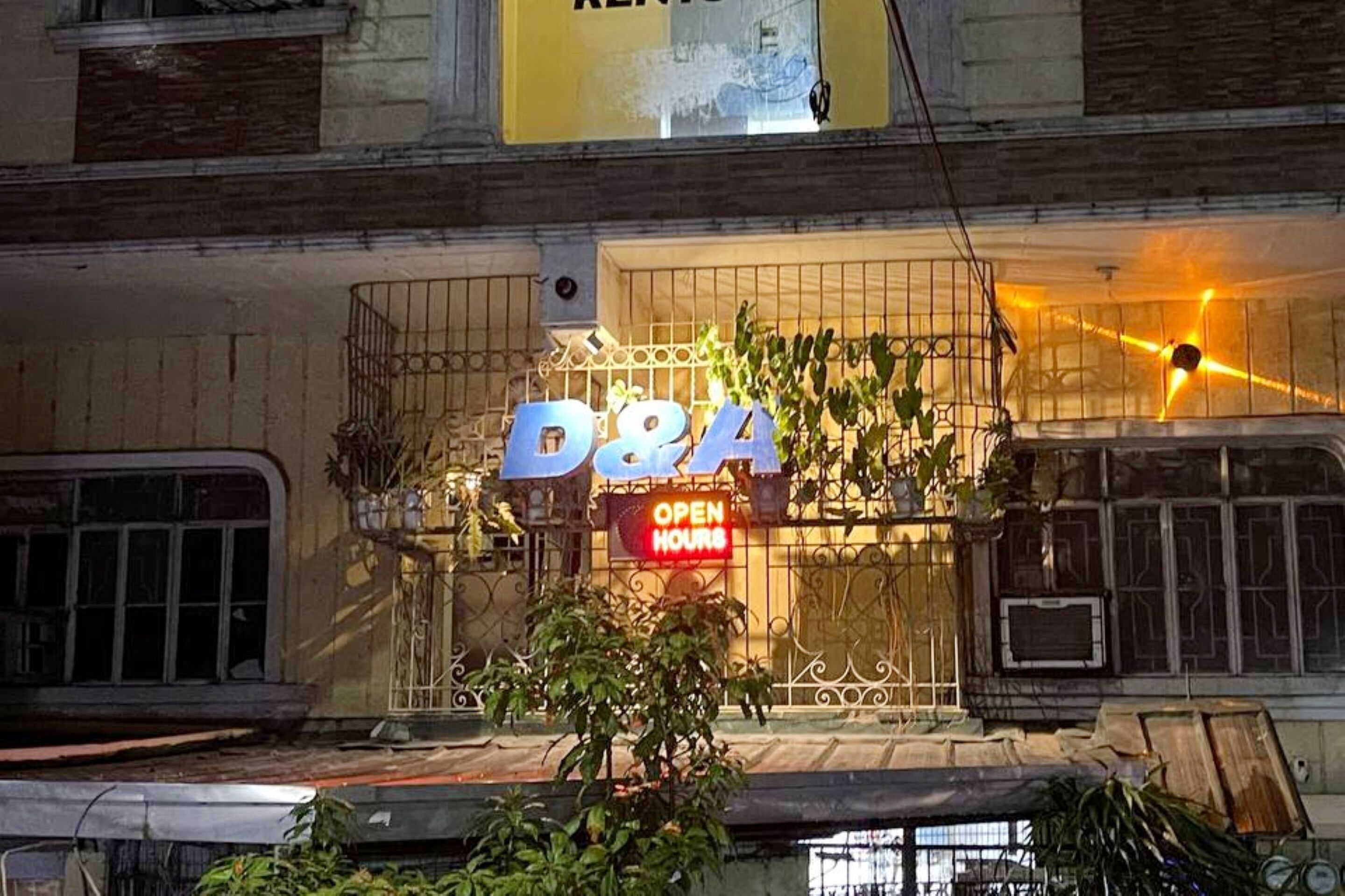Un magasin D&A avec une enseigne indiquant les « HORAIRES D’OUVERTURE », entouré de plantes et d’une clôture en métal.