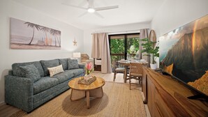 A living room with a sofa, coffee table, and TV. There is a ceiling fan, a window with curtains, and a painting on the wall. - BRAND NEW REMODELED 2/2 Family Getaway! Walk to Amazing Beach, AC & WiFi (Lihue)