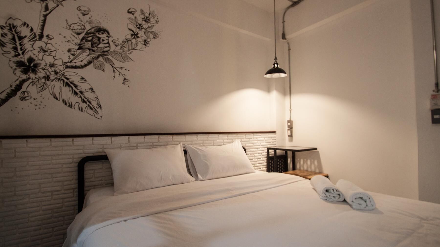 Uma cama bem arrumada com lençóis brancos, cabeceira preta e um mural de aves e flores na parede.