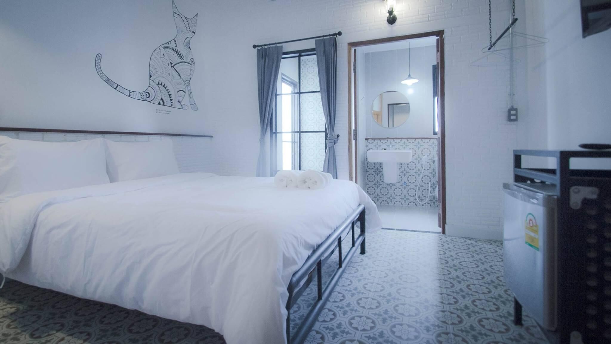 A bedroom with a bed, a wall-mounted cat illustration, a window with curtains, a bathroom with a towel rack, and patterned flooring.