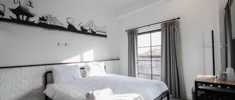 A hotel room with a bed, white linens, a window with curtains, and a wall with a black and white mural.