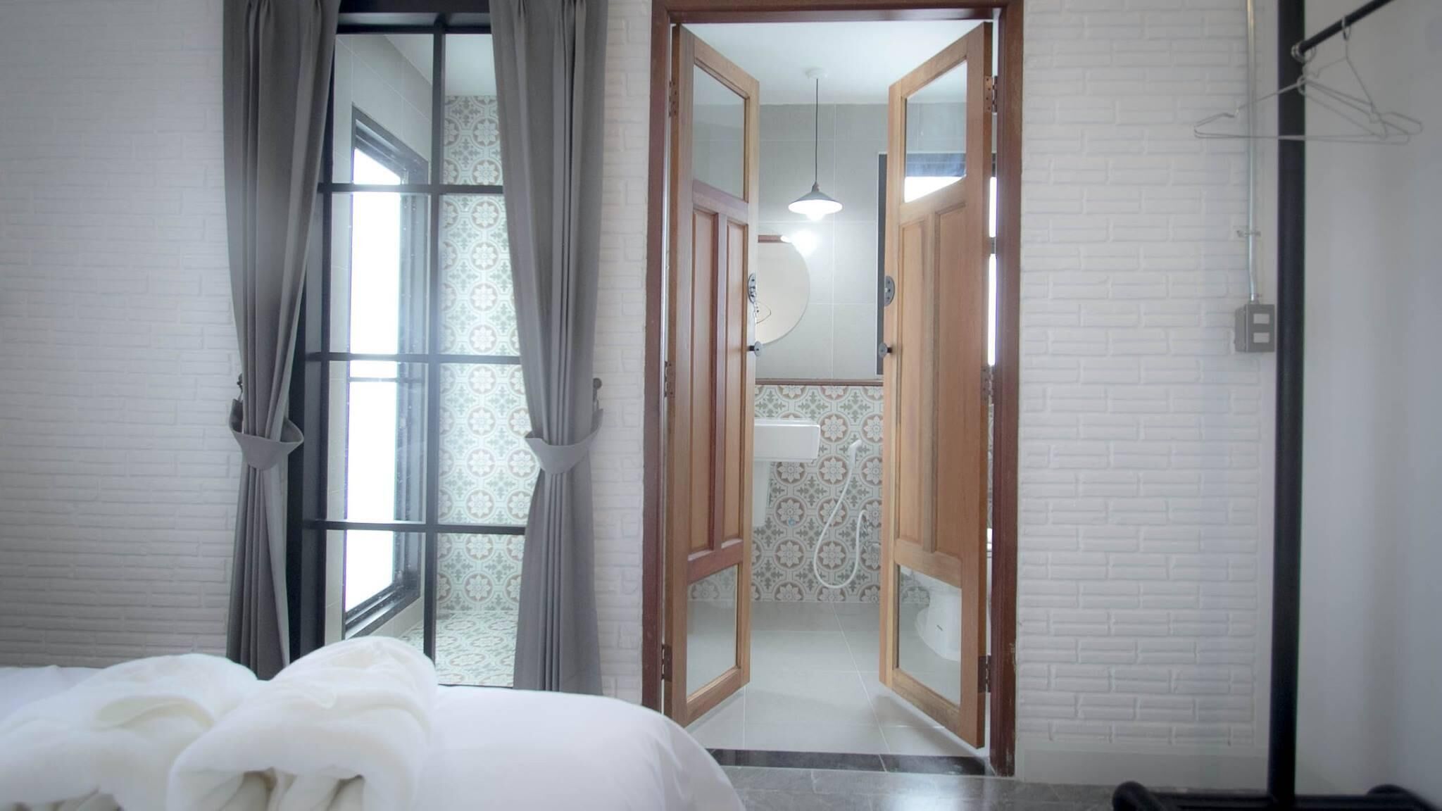 A hotel room with a bed, a window with curtains, a wooden door leading to a bathroom, and a hanging light fixture.