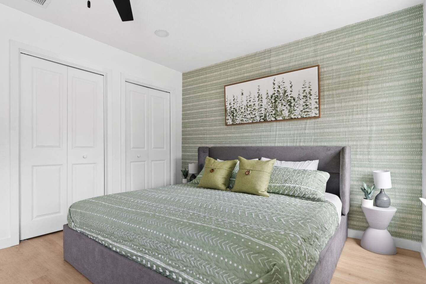 A bedroom with a large bed, a grey headboard, and a green patterned bedspread. There is a white door on the left and a wall with a green patterned wallpaper.