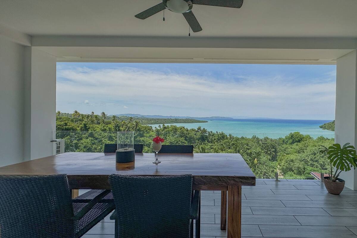 A dining area with a table and chairs, a ceiling fan, and a view of a tropical landscape with a body of water.