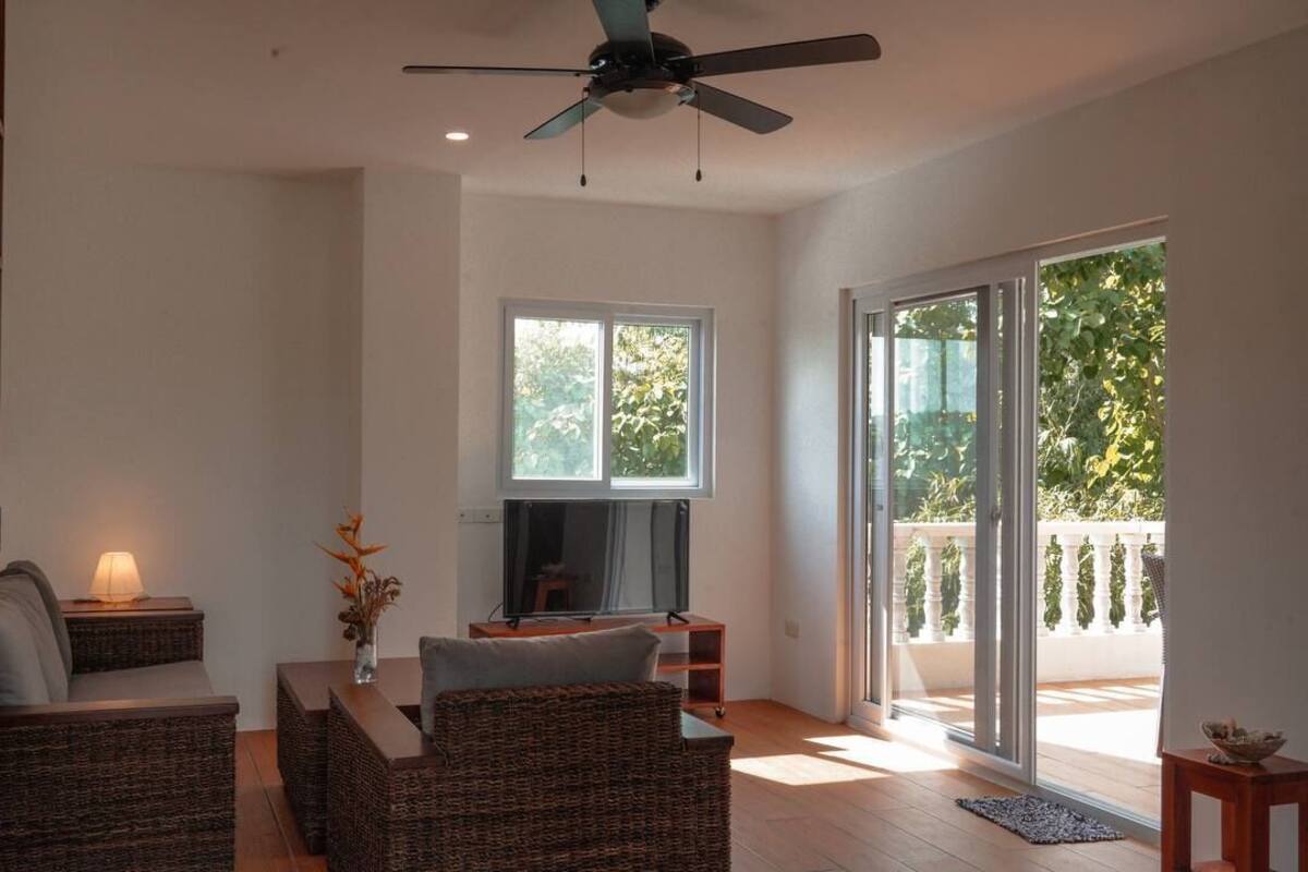 A spacious living room with a large TV, wicker furniture, a ceiling fan, and a sliding glass door leading to a balcony.