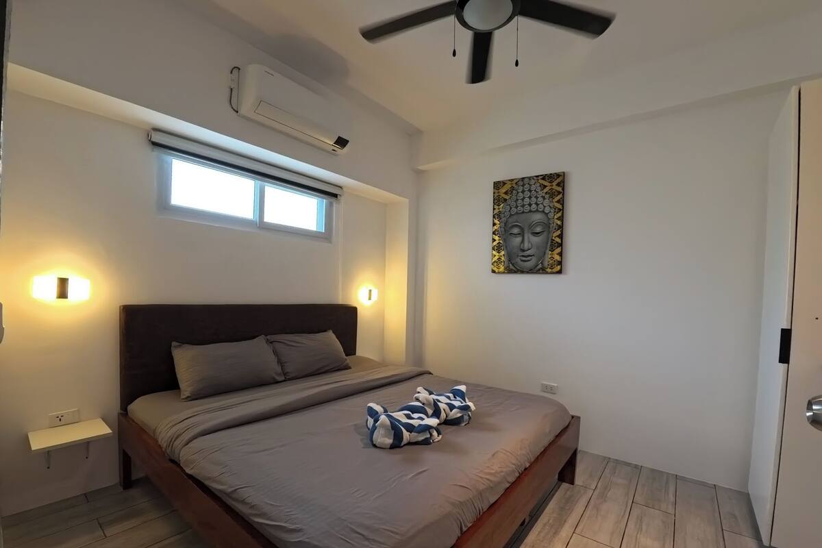 A bedroom with a bed, bedside tables, an air conditioning unit, a ceiling fan, a window, and a wall-mounted artwork.