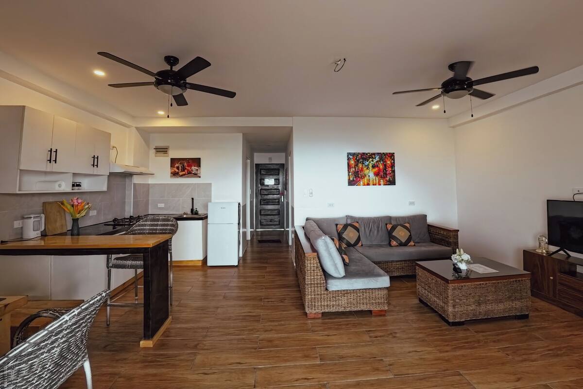 A modern apartment with a kitchen, dining area, and living room. It includes two ceiling fans, a sofa, a coffee table, and a TV.