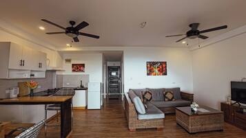A modern apartment with a kitchen, dining area, and living room. It includes two ceiling fans, a sofa, a coffee table, and a TV.
