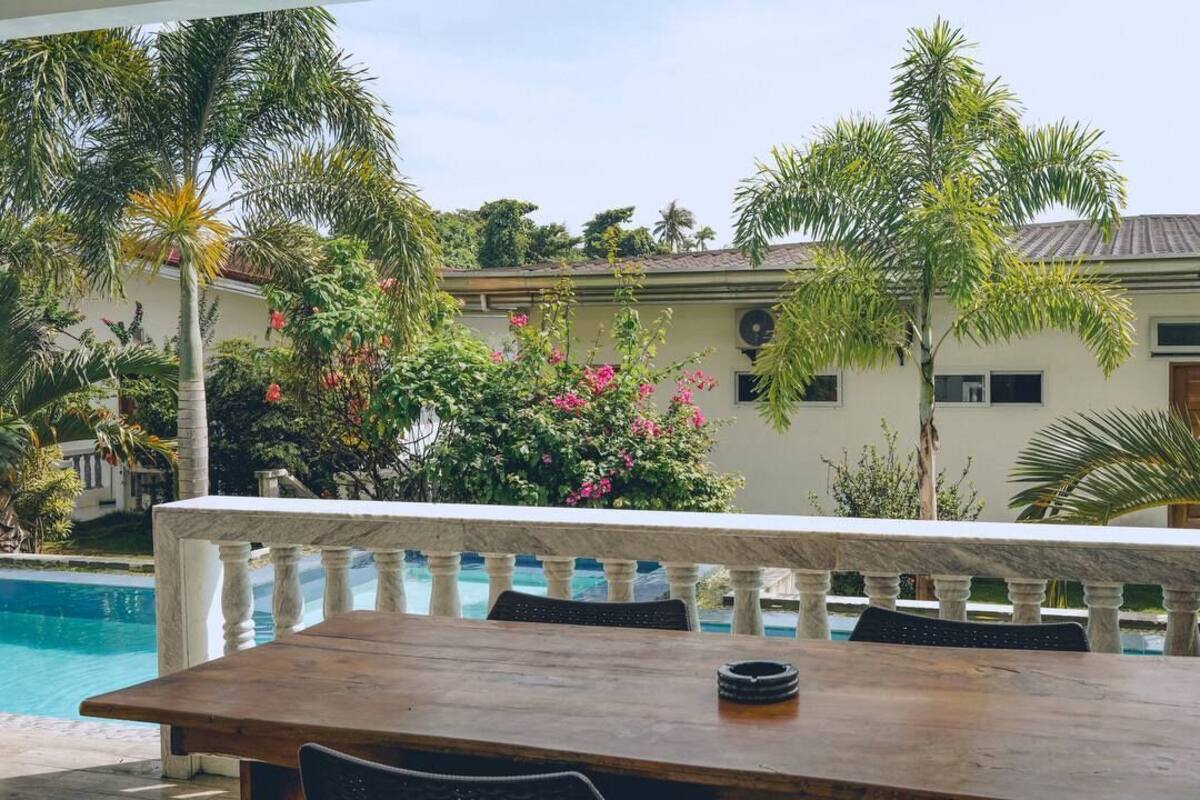 A poolside area with a wooden table and chairs, a stone balustrade, and a building with a tiled roof in the background.