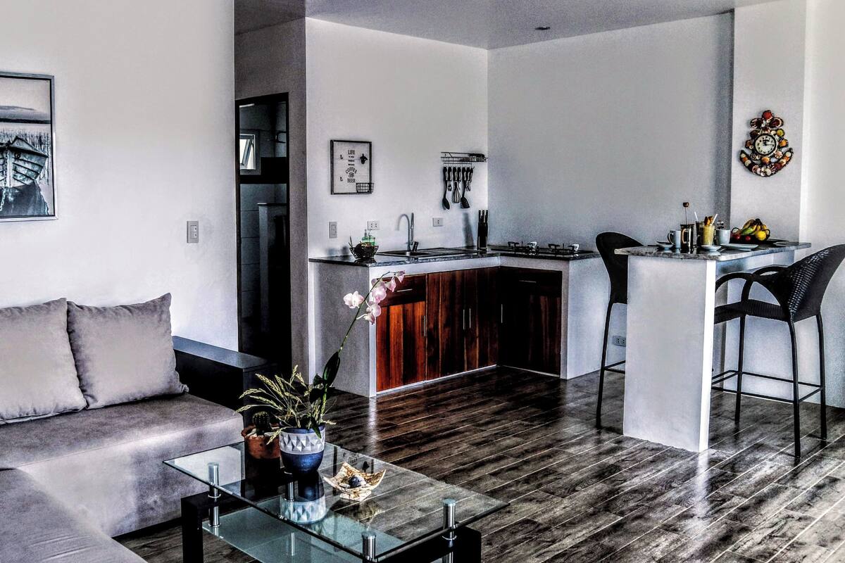 A modern living room with a grey sofa, a glass coffee table, and a kitchen area with a white countertop and dark wooden cabinets.