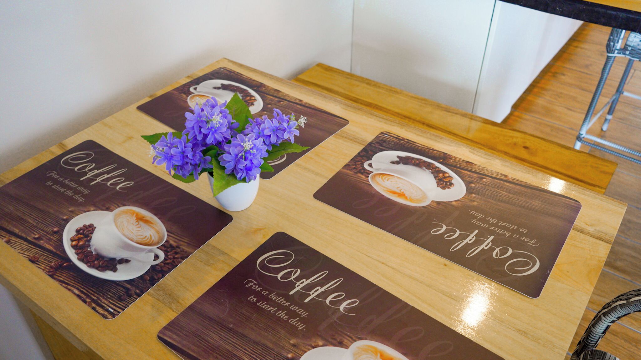 A wooden table with four placemats featuring coffee-themed designs and a vase with purple flowers.