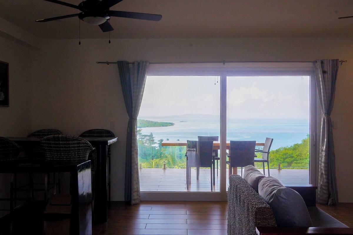 A living room with a view of the sea, a ceiling fan, and a dining area with chairs and a table.