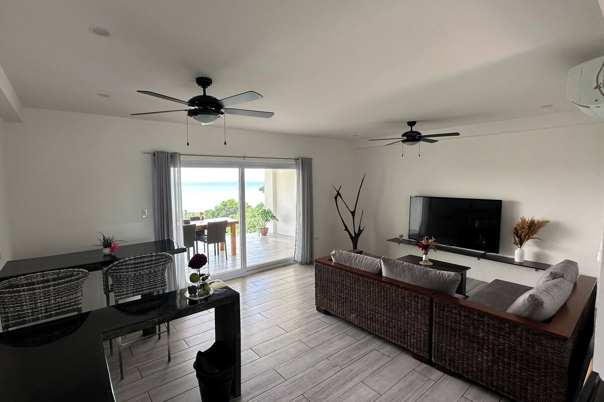 A modern living room with a dining area, two ceiling fans, a flat-screen TV, and a large sliding glass door leading to a balcony with a view.