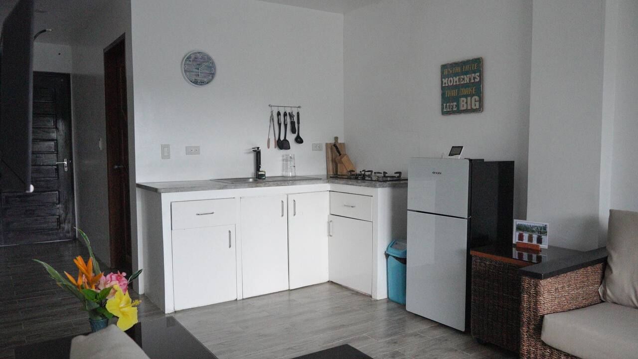 Une cuisine moderne avec des armoires blanches, une cuisinière encastrée et un réfrigérateur. Il y a une horloge au mur et un panneau affichant une citation inspirante.