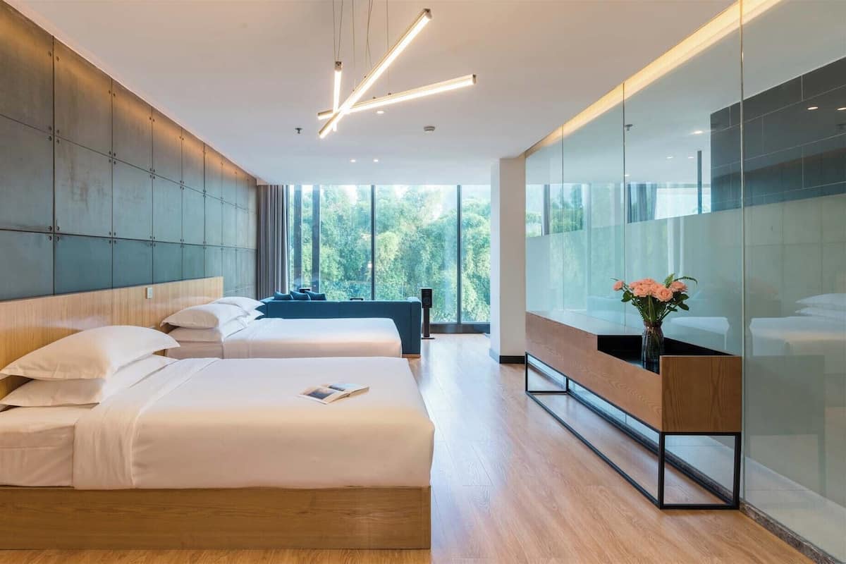 Una habitación de hotel moderna con dos camas, un ventanal con vistas a árboles y un separador de vidrio con un florero.