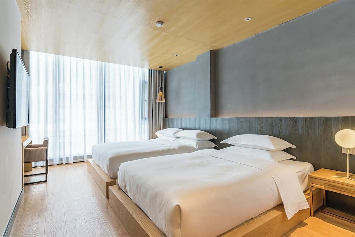Una habitación de hotel moderna con dos camas, un televisor montado en la pared, una mesita de noche con lámpara y un ventanal con cortinas.