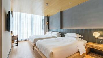 A modern hotel room with two beds, a TV mounted on the wall, a bedside table with a lamp, and a large window with curtains.