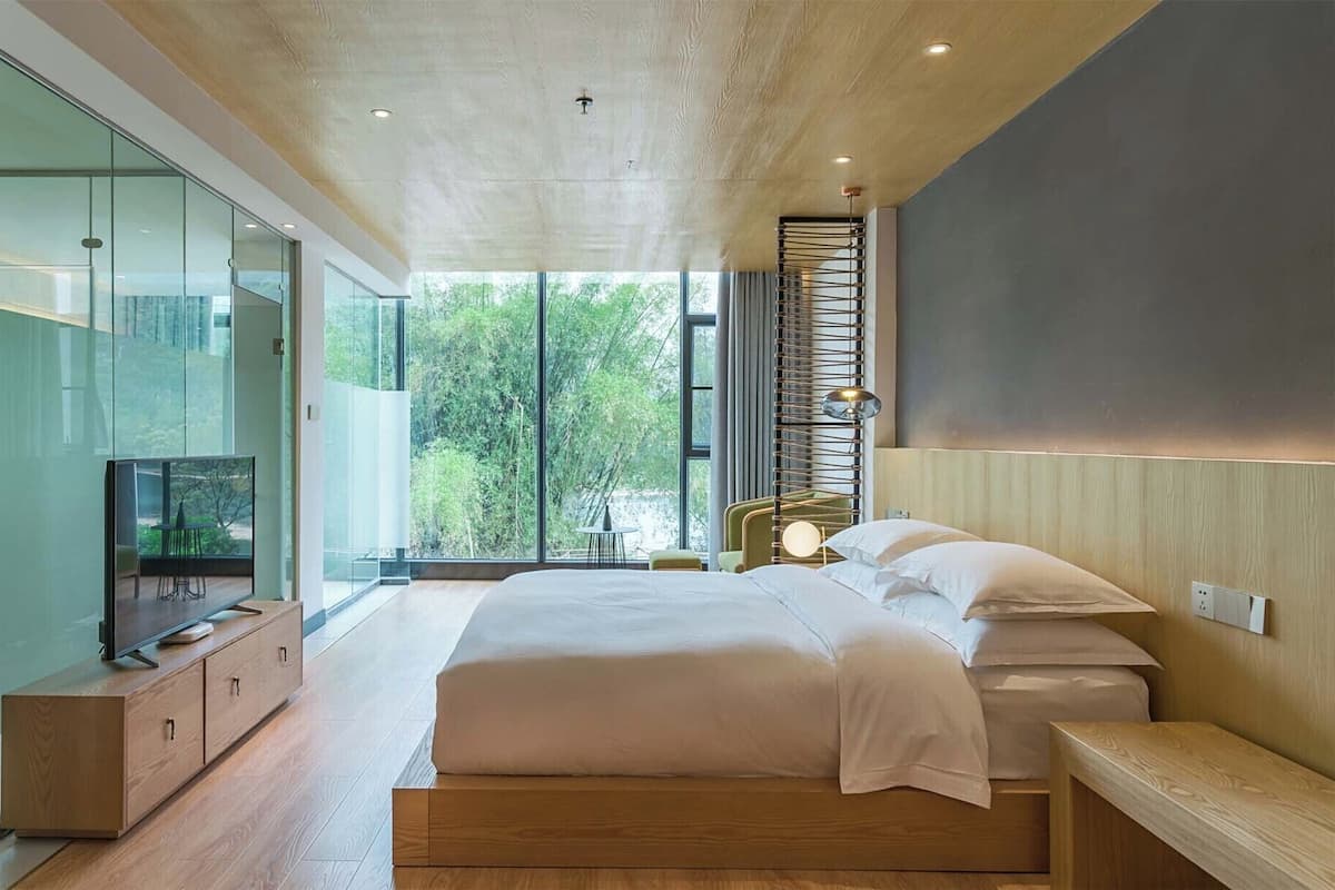 Un dormitorio moderno con una cama grande, un televisor sobre un mueble de madera y vistas a la vegetación a través de amplias ventanas.