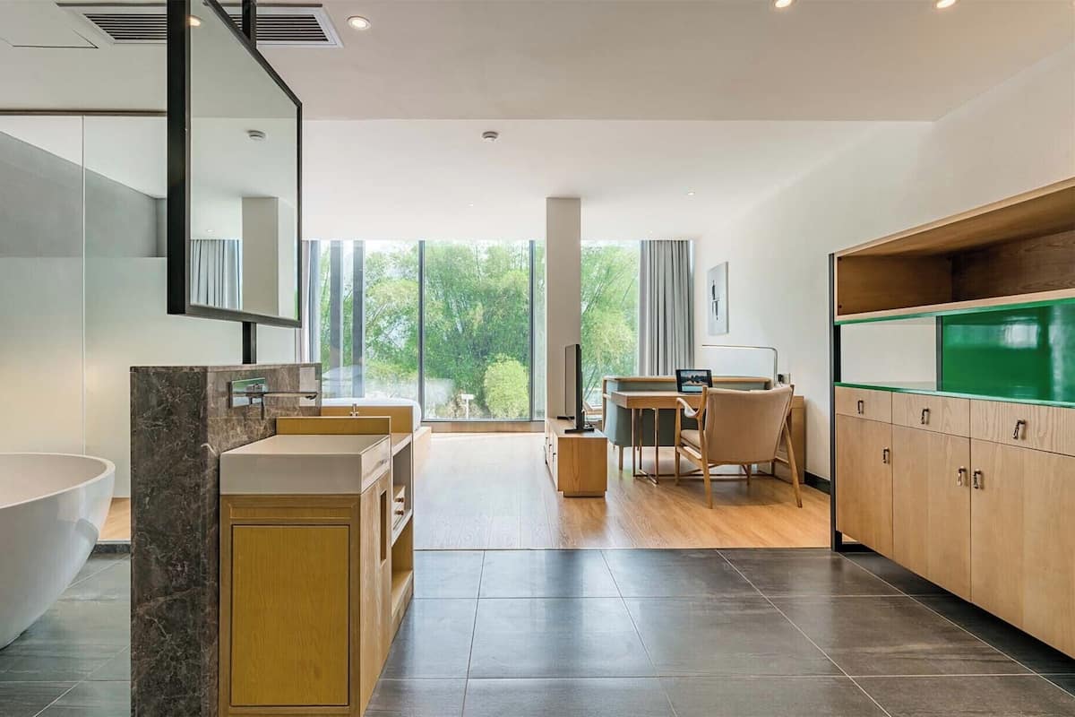 Un baño moderno con una bañera grande, un tocador con espejo y una zona de ducha. Hay una sala de estar con una mesa y una silla, y vistas a zonas verdes.