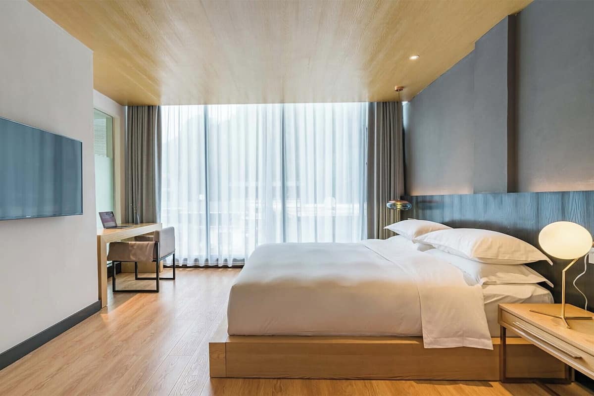 Habitación de hotel moderna con una cama grande, un escritorio con una computadora portátil, un televisor montado en la pared y un ventanal con cortinas.