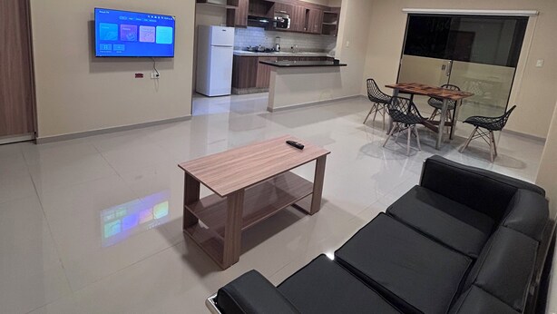 Een moderne woonkamer met een leren zwarte bank, een houten salontafel en een flatscreen televisie aan de muur.