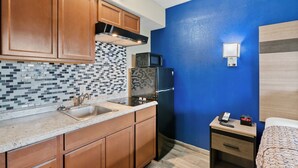 A compact kitchen with wooden cabinets, a tiled backsplash, a stainless steel sink, a microwave, and a refrigerator. There's a small bedside table with a phone and a digital clock. - 2nd level pool front • Kitchen • Walk to Beach (Rockport)