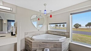 A modern bathroom with a large bathtub, a heart-shaped mirror, and a view of palm trees through a window. - Waterview King Suite • Pool front, ground level (Rockport)