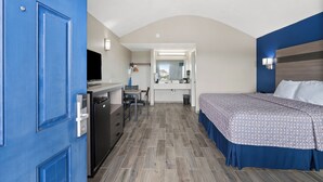 Une chambre d’hôtel compacte avec une porte bleue, un lit recouvert d’une couvre-lit à motifs, une petite kitchenette, un bureau avec une chaise et un miroir au-dessus du lavabo.