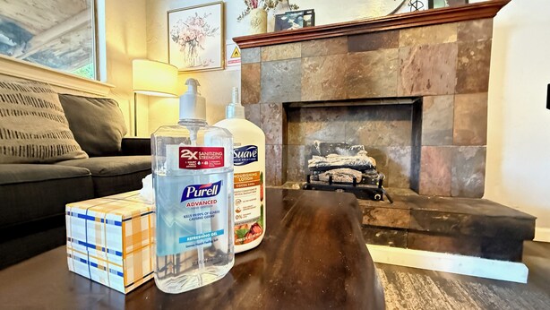Een woonkamer met een open haard, een bank en een tafel waarop desinfecterende handgel en lotion staan.
