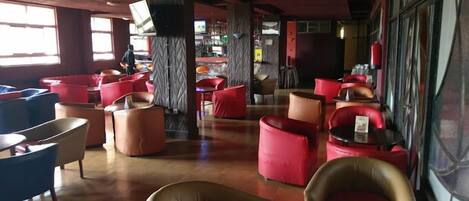 A modern lounge area with red and beige seating, a bar in the background, and large windows providing natural light.