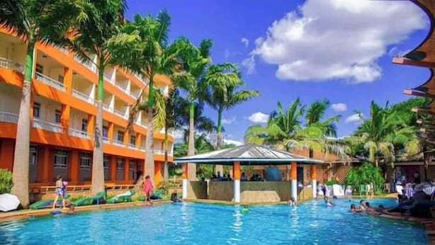 A hotel pool area with a clear blue pool, palm trees, and a multi-story building with orange and white exterior walls.
