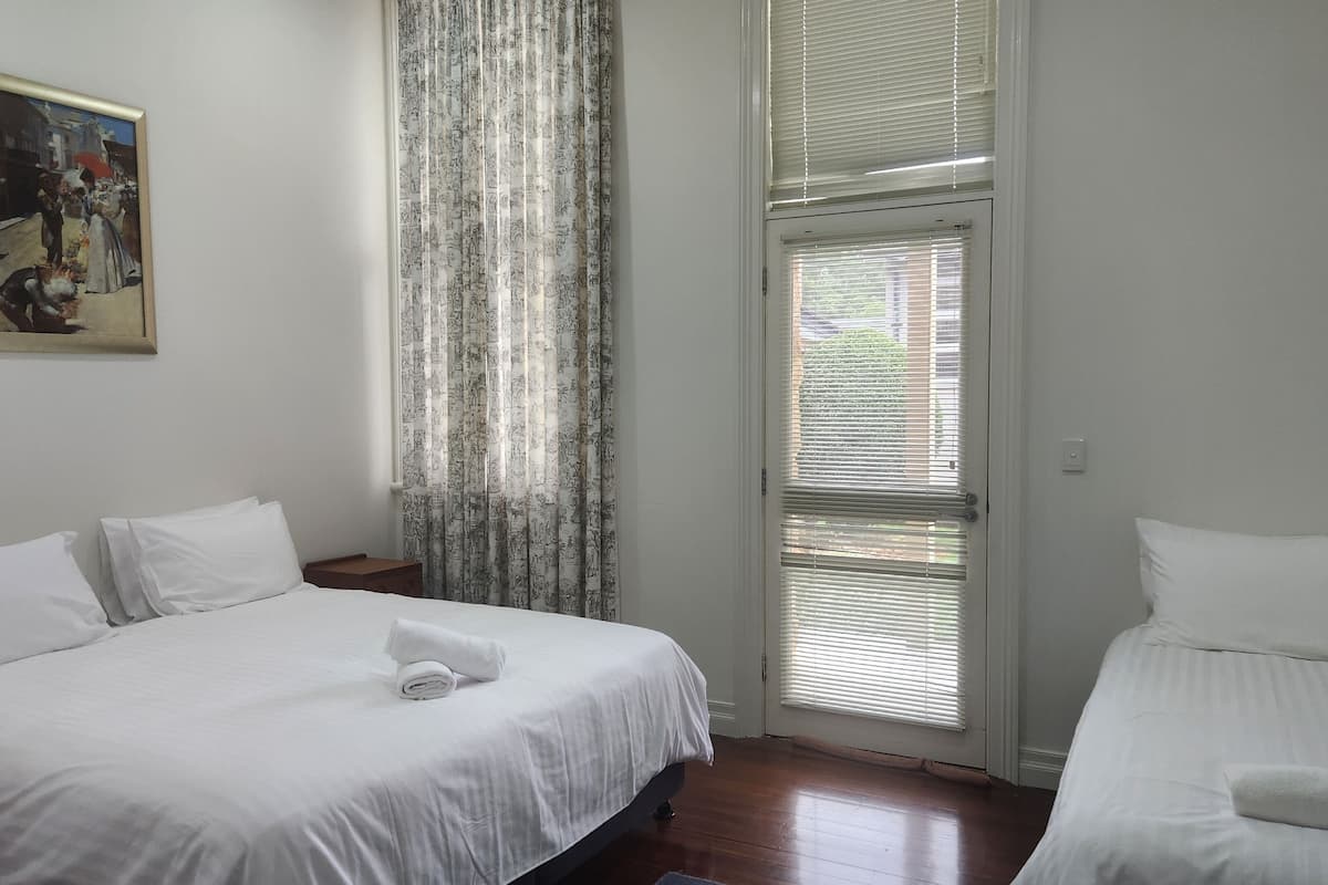 A hotel room with two beds, a painting on the wall, and a window with blinds.