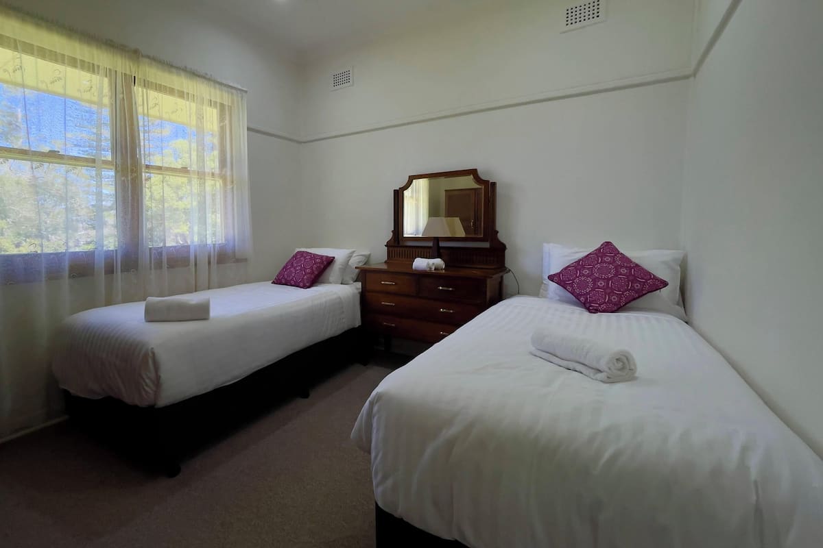 A hotel room with two single beds, a wooden dresser, a mirror, and a window with sheer curtains.