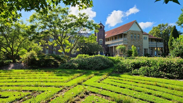 Un edificio grande con una torre, rodeado de abundante vegetación y un jardín cuidado.
