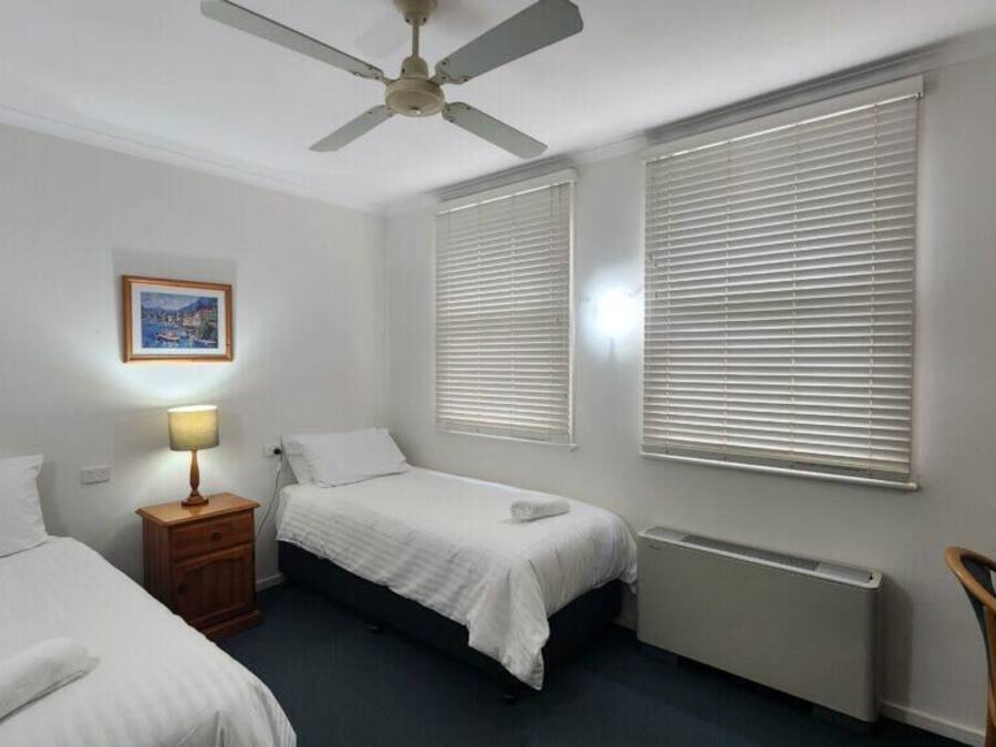 A hotel room with two single beds, a ceiling fan, a nightstand with a lamp, a radiator, and a chair.