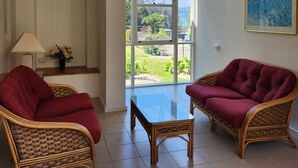 Una habitación con dos sillones de mimbre, una mesita, una lámpara, un florero con flores y un ventanal con vistas a zonas verdes y edificios.