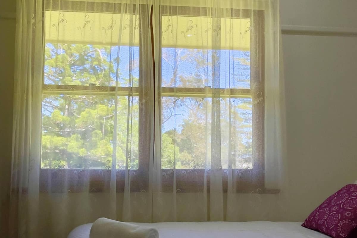A bed with a white sheet and a rolled towel, a purple pillow, and a window with sheer curtains.