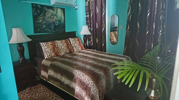 Een slaapkamer met een bed, twee bedlampen, een bedspreide met luipaardprint, een grote spiegel en een kamerplant.