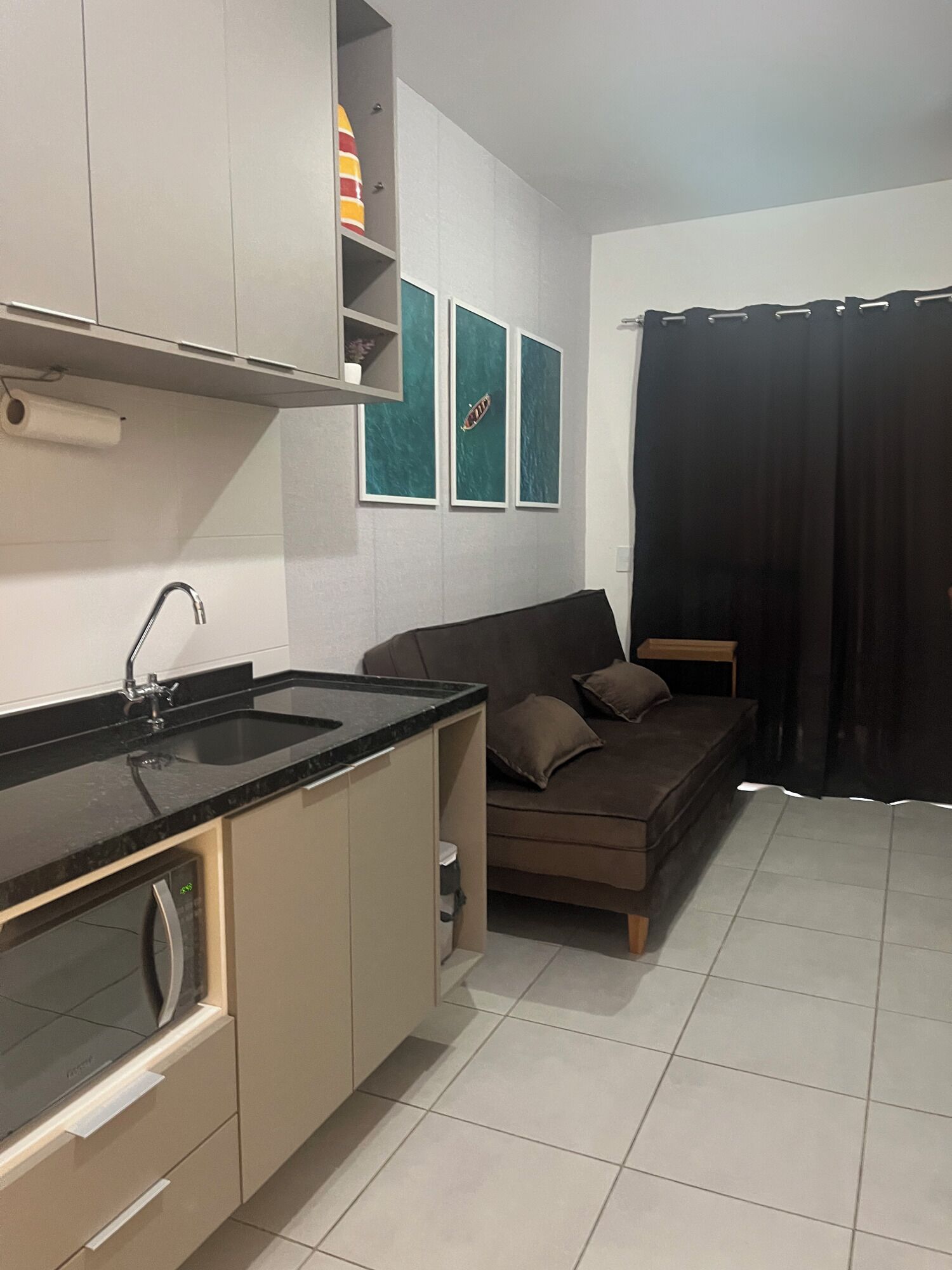 Uma cozinha compacta com bancada preta, forno embutido e pia. Um sofá marrom está posicionado contra a parede, e há três quadros pendurados acima.