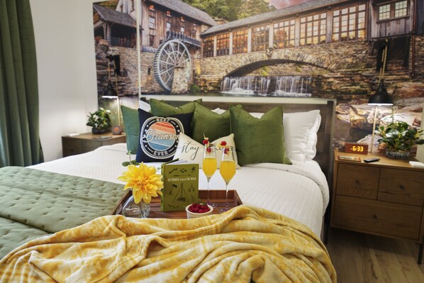 A hotel room with a bed, a nightstand, a tray with drinks, a book, and a painting of a waterfall.