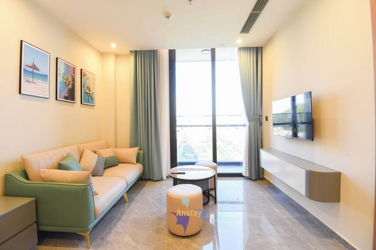 Una habitación de hotel moderna con sofá, una mesita redonda, televisor montado en la pared y un balcón con cortinas.
