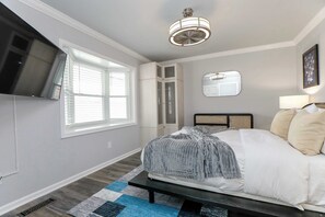 Ein Schlafzimmer mit einem Bett, einem an der Wand montierten Fernseher, einem Fenster mit Jalousien, einem Deckenventilator und einem Spiegel.