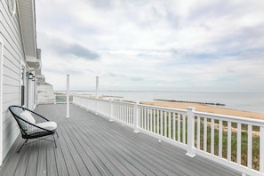 A wooden deck with a white railing, a black chair, and a view of the beach and ocean.
