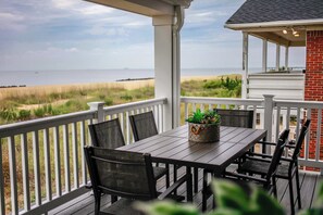 A covered patio with a wooden table and chairs, overlooking a beach and dunes.