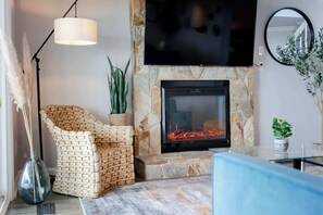 Una sala de estar con chimenea de piedra, un televisor de pantalla plana montado encima y una silla tejida, además de una planta en maceta.